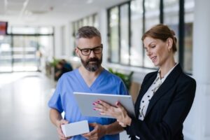 A healthcare administrator shows a healthcare provider a tablet while standing in a hospital corridor.
