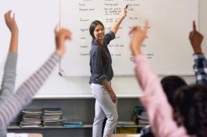A bilingual teacher standing by a whiteboard in front of students raising their hands.