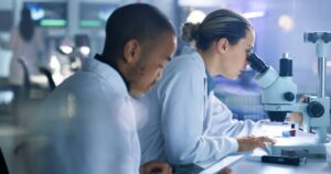Forensic science technicians view crime scene evidence under a microscope in a lab.