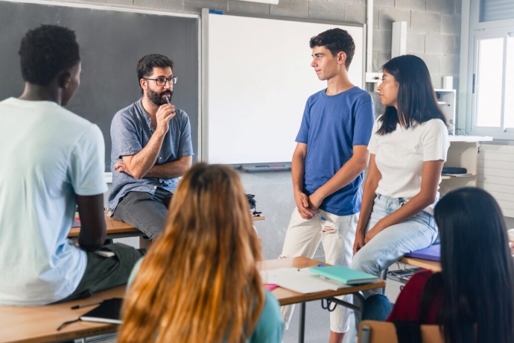 A high school teacher leaning against his desk talks to a group of students.