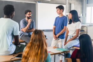 A high school teacher leaning against his desk talks to a group of students.